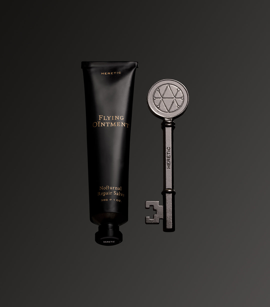 A black squeeze tube labeled 'FLEYING OINTMENT' stands beside a decorative silver key engraved with 'Heretic' and a geometric symbol, on a dark gray gradient background.