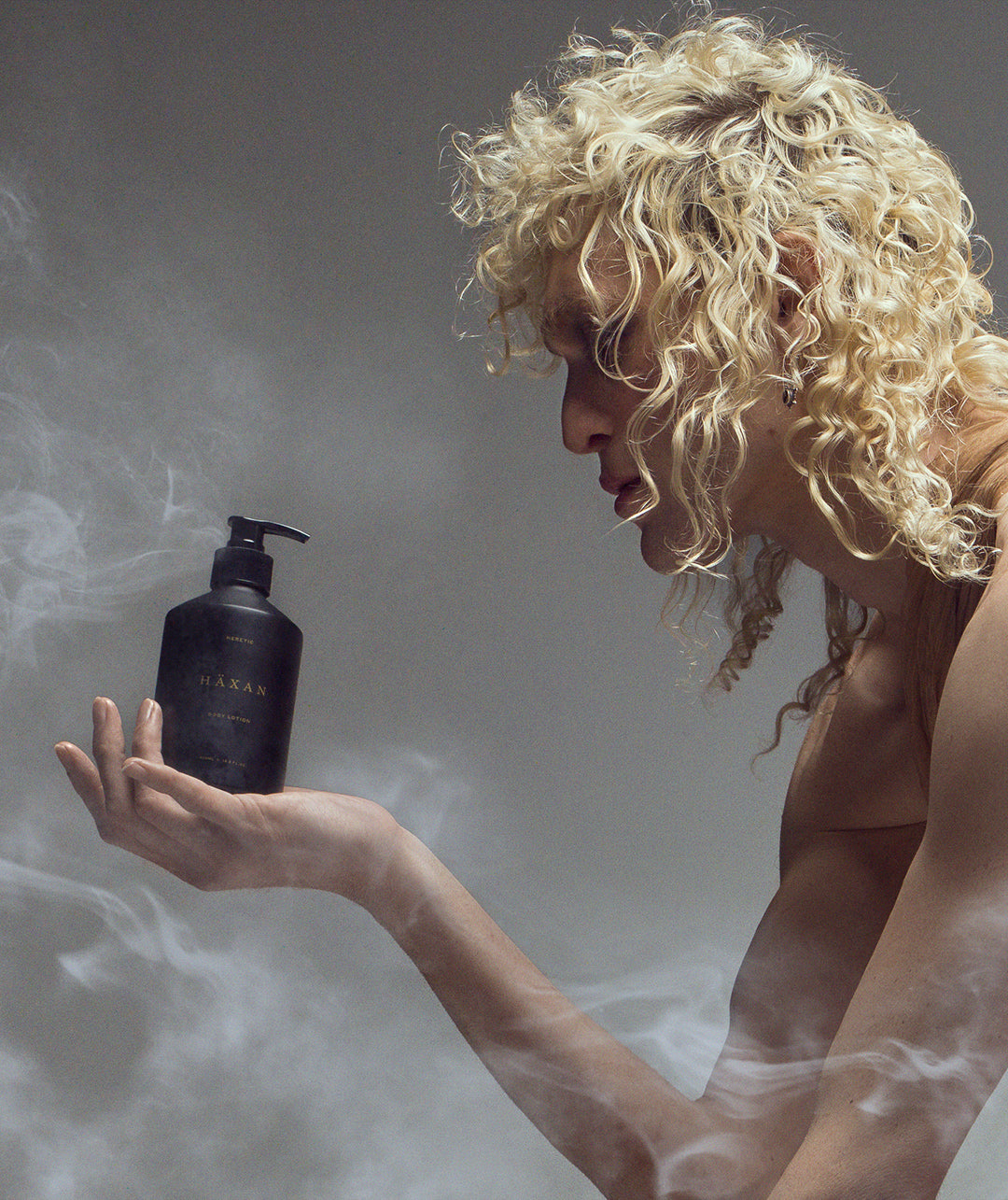 A model with voluminous curly blonde hair holds a matte black pump bottle labeled 'Haxan' in their hand, set against a smoky gray studio background.